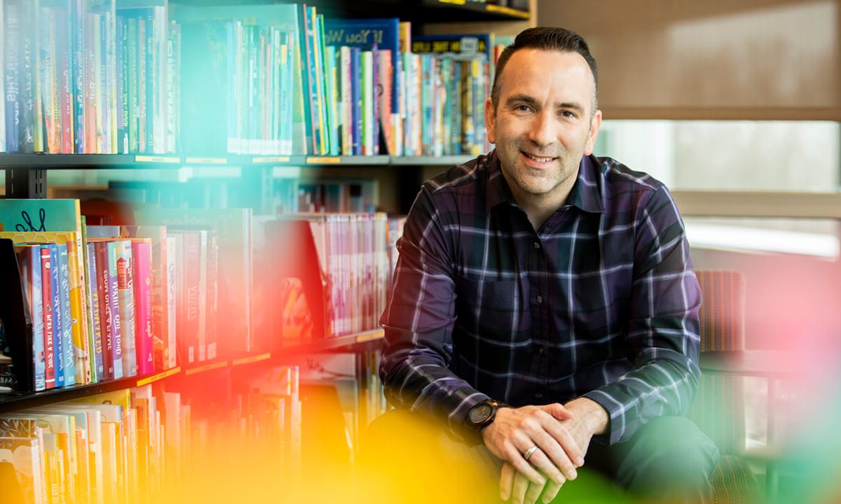 Man looks toward camera smiling. There is a library bookshelf behind him.